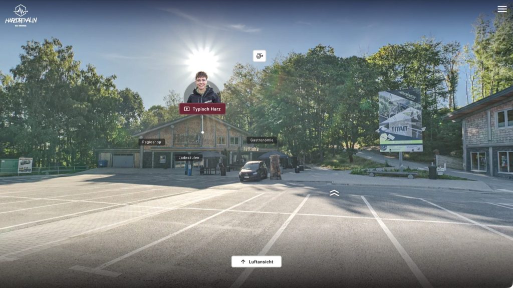 Screenshot of the tour looking across a parking lot toward the main buildings, with labeled hotspots for amenities (Regioshop, Gastronomie, Ladesäulen) and a video hotspot “Typisch Harz,” plus a “Luftansicht” (aerial view) toggle.